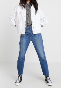 Giacca di jeans bianca, camicia a righe bianco e nero, jeans blu a vita alta, sneakers nere, in piedi con le mani sui fianchi, look casual.