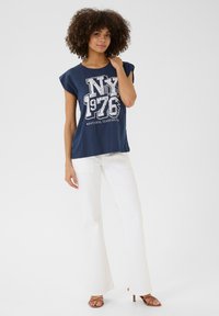 Jūras spēku t-krekls ar retro baltu grafisku printu, īsām piedurknēm, savienots ar baltām flared biksēm un brūniem augstpapēžu zandaliem.