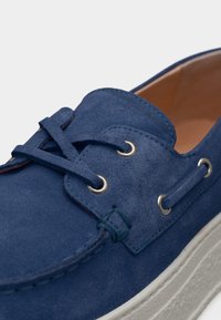 Μπλε σουέτ παπούτσι σκάφους με χοντρή λευκή λαστιχένια σόλα, μεταλλικά οπτικά και ταιριαστά μπλε κορδόνια σε απλό ανοιχτό φόντο.