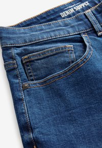 Blauwe denim jeans met voorzak, koperen stiksels, riemlus, klinknagel en "Denim Supply" label op de tailleband.