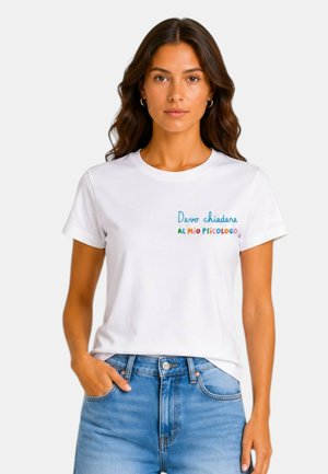 Donna con lunghi capelli mossi che indossa una t-shirt bianca con testo italiano colorato e jeans blu, in piedi con una mano in tasca.