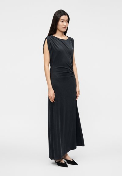 Woman with long dark hair wears a sleeveless black maxi dress and black pointed-toe heels, standing against a plain white background.