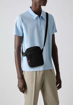 Man wearing light blue short-sleeve polo and beige trousers carrying a small black crossbody bag with red zipper pulls.