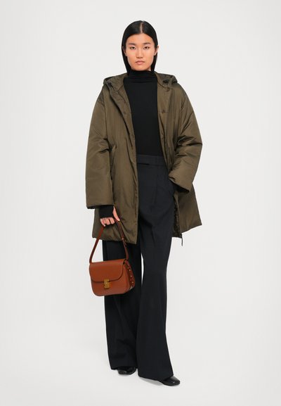 Frau trägt einen olivgrünen oversized Mantel, einen schwarzen Rollkragenpullover, weite schwarze Hosen, schwarze Schuhe und hält eine braune Lederhandtasche.