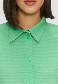 Blusa di un verde menta con texture e colletto. Presenta chiusura con bottoni, cuciture a vista e un tessuto leggero leggermente trasparente.