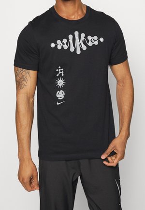 Homme portant un t-shirt noir Nike avec une onde sonore blanche abstraite et des icônes sportives verticales sur le devant, tenant l'ourlet du t-shirt de la main droite.