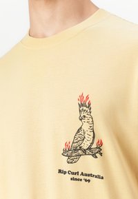 Beige t-shirt med sort og rød grafik af en fugl med flammende krone, der sidder på en brændende gren, med teksten "Rip Curl Australia siden '69" nedenunder.