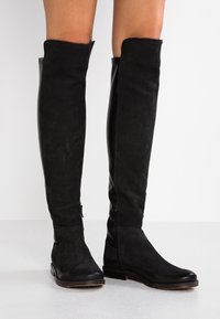 Botas altas negras de muslo, hechas de ante y cuero, con una textura suave, cremalleras laterales y suela plana. Diseño minimalista.