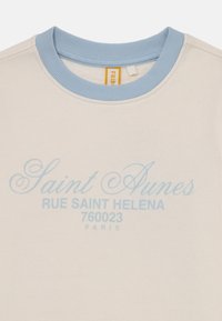 Lys beige sweatshirt med blå ribbet krage, med trykt tekst: "Saint Aunes," "RUE SAINT HELENA," og "760023 PARIS" i lys blå.