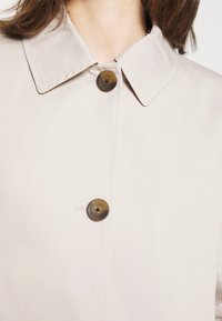 Beige jacka med spetsig krage, två bruna knappar och synlig sömnad. Slät tyg med matt yta, utan mönster eller detaljer.