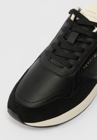 Sneaker din piele neagră și suede cu șireturi negre, talpă albă și marcaj auriu "Tommy Hilfiger" pe lateral și pe limba pantofului.