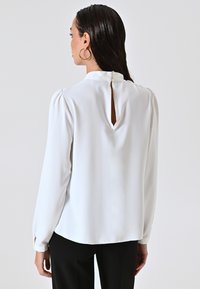 Blusa bianca a maniche lunghe con colletto alto, apertura a chiave sul retro e chiusura a bottone. Tessuto morbido con una texture liscia.