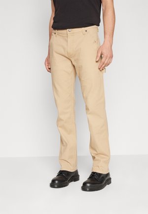 Mann trägt beige gerade geschnittene Hose, schwarzen Gürtel, schwarzes Hemd und schwarze Schnürschuhe, steht vor einem schlichten weißen Hintergrund.
