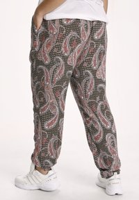 Braun gemusterte Hose mit einem Paisleymuster in Rot und Blau, ausgestattet mit einem elastischen Bund und seitlichen Taschen, kombiniert mit weißen Turnschuhen.