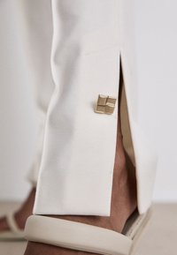 Hellbeige, maßgeschneiderte Hosen verfügen über einen scharfen seitlichen Schlitz mit einem subtilen goldenen Akzent am Saum und werden mit einem hellen Sandalen kombiniert.