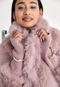 Femme aux cheveux foncés portant un manteau rose duveteux et des manches en maille assorties, avec des bracelets argentés et dorés, tenant le col du manteau.