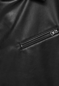 Matière en cuir noir avec une poche zippée horizontale fermée équipée d'une languette en métal et de bords cousus.