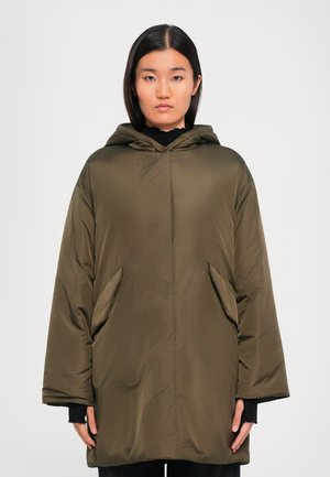 Woman with straight dark hair wearing an oversized olive green hooded puffer jacket with large front pockets, standing against a light background.