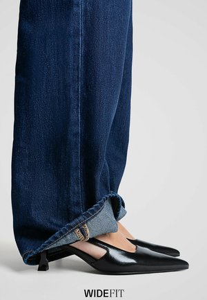 Black patent leather slip-on shoes with a pointed toe and cut-out design, paired with dark blue cuffed denim jeans.