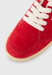 Baskets en daim rouge avec des lacets crème, dotées d'un bout arrondi et d'une semelle en caoutchouc beige. Texture lisse et détails cousus visibles.
