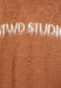 Hnědý chlupatý textil se bílým nápisem "TWO STUDIOS." Materiál se zdá být jemný s huňatou strukturou.