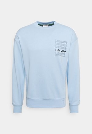 Jasnoniebieska bluza z długim rękawem, ze ściągaczami przy rękawach i u dołu oraz napisem „Lacoste” powtórzonym pięć razy na lewej piersi w różnych odcieniach.