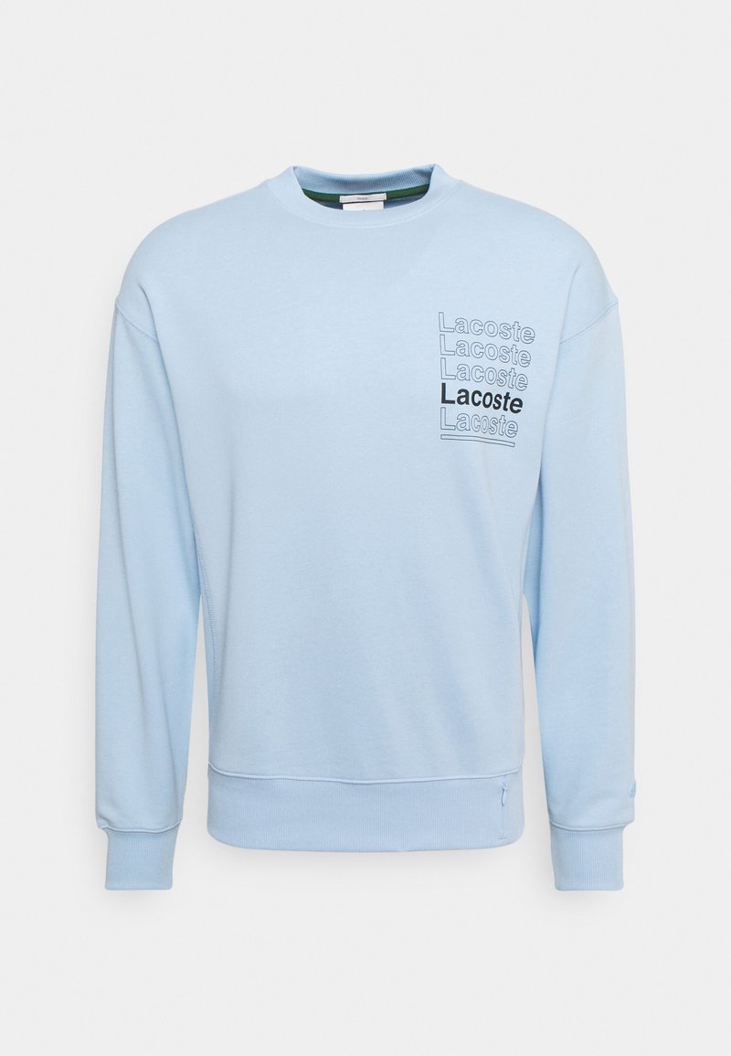 Sweat-shirt bleu clair à manches longues avec poignets et ourlet côtelés, et « Lacoste » répété cinq fois sur le côté gauche de la poitrine dans différentes nuances.