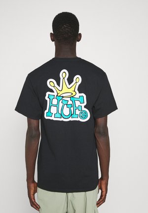 Homme portant un t-shirt noir avec un grand texte coloré "HUF" et un motif de couronne jaune dans le dos, debout devant un fond clair et uni.