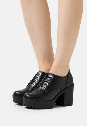 Scarpe piattaforma nere con lacci, texture a rilievo effetto coccodrillo, punta rotonda e tacco spesso. Presentano occhielli in argento e un design elegante.