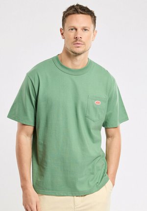 Homme aux cheveux courts portant un t-shirt vert à manches courtes avec une poche poitrine et un pantalon beige, debout devant un fond clair uni.