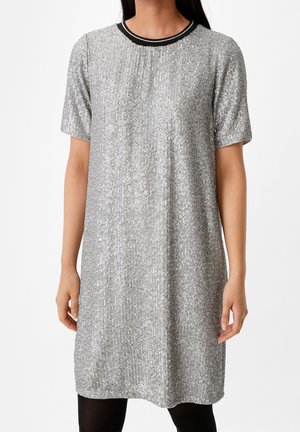 Frau trägt ein silbernes Paillettenkleid knielanger Länge mit kurzen Ärmeln und einem schwarzen, runden Ausschnitt, steht vor weißem Hintergrund.