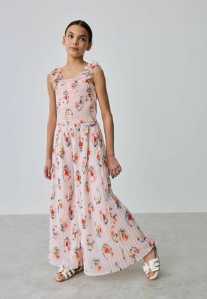 Fată care poartă o rochie lungă roz cu flori fără mâneci, cu umeri cu volane și sandale albe, stând pe o podea gri, în fața unui perete simplu.