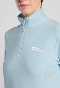Pullover in pile azzurro chiaro con colletto a mezza zip, caratterizzato dal logo "Jack Wolfskin" ricamato in bianco sul lato sinistro del petto.