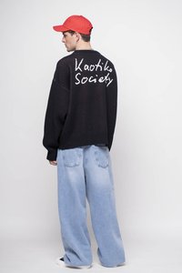 Crna oversized sweatshirt s bijelim natpisom "Kaotiko Society" na leđima, uparen s svijetloplavim širokim trapericama. Crvena kapa i tenisice upotpunjuju izgled.