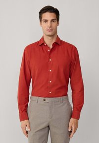 Rotes Hemd mit langen Ärmeln, weicher Textur und weißen Knöpfen; kombiniert mit hellgrauen karierten Hosen.