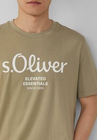 Olijfgroene t-shirt van katoen met witte tekst die leest "s.Oliver ELEVATED ESSENTIALS SINCE 1969" in een casual lettertype.