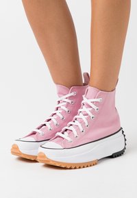 High-top sneakers in light pink canvas with white laces, black and white sole, and a textured orange outsole. Features a pull tab at the back.