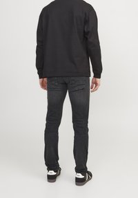 Camisa negra de manga larga, jeans oscuros y zapatos negros con detalles en blanco, vistos desde atrás, destacando líneas limpias y un buen ajuste.