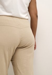Pantalons beiges légers avec une texture subtile, dotés d'une taille élastique et de détails de couture, présentés de dos.