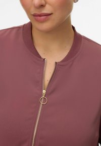 Bordeauxrode bomberjack met geribde kraag en gouden ritssluiting. Gladde stoftextuur, voorzien van een ronde ritstrekker voor gemakkelijke toegang.