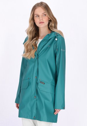 Donna che indossa un impermeabile con cappuccio verde acqua con tasche anteriori, in piedi contro uno sfondo bianco, che guarda di lato.