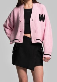 Růžový pletený kardigan s žebrovanou texturou, s černou nášivkou ve tvaru "W", zapínáním na knoflíky a kontrastním černým lemováním. Kombinováno s černou sukní.