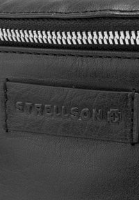 Detal na czarnej skórzanej torbie z widocznym srebrnym zamkiem błyskawicznym oraz wytłoczonym napisem "STRELLSON" i logo na teksturowanej powierzchni.