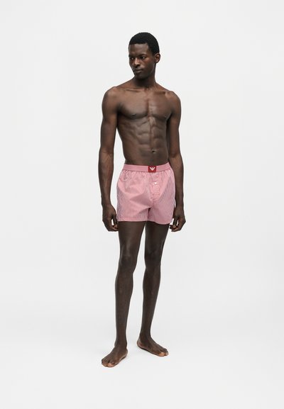 Άνδρας μοντέλο με σκουρόχρωμο δέρμα, φορώντας κόκκινα και λευκά ριγέ μποξεράκια, στέκεται ξυπόλητος σε ένα απλό ανοιχτό γκρι φόντο.