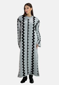 Lang grå strikket kjole med sorte zigzag-mønstre, brede ærmer og rund halsudskæring, kombineret med sorte ankelstøvler.