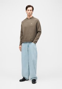 Mees, kes kannab pruuni kampsunit ja laiu helerohelisi pükse koos mustade kingadega, seisab ühevärvilise valge tausta ees.