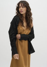 Schwarze, oversized Button-Up-Bluse, die über einem seidigen, senfgelben Slip-Kleid mit V-Ausschnitt getragen wird. Lockige Haare, neutraler Hintergrund.
