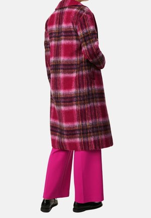 Personne portant un long manteau à carreaux rouge et violet avec un pantalon rose vif et des chaussures noires, debout les mains dans les poches du manteau.