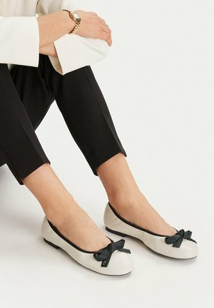 Mujer con bailarinas blancas con lazos negros, pantalones negros recortados y un reloj de pulsera dorado, sentada con las piernas cruzadas.