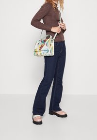 Multikleurige handtas met bloemenpatroon op een mintgroene basis, voorzien van een platte klep en een verstelbare band. Draagt bij donkere jeans.